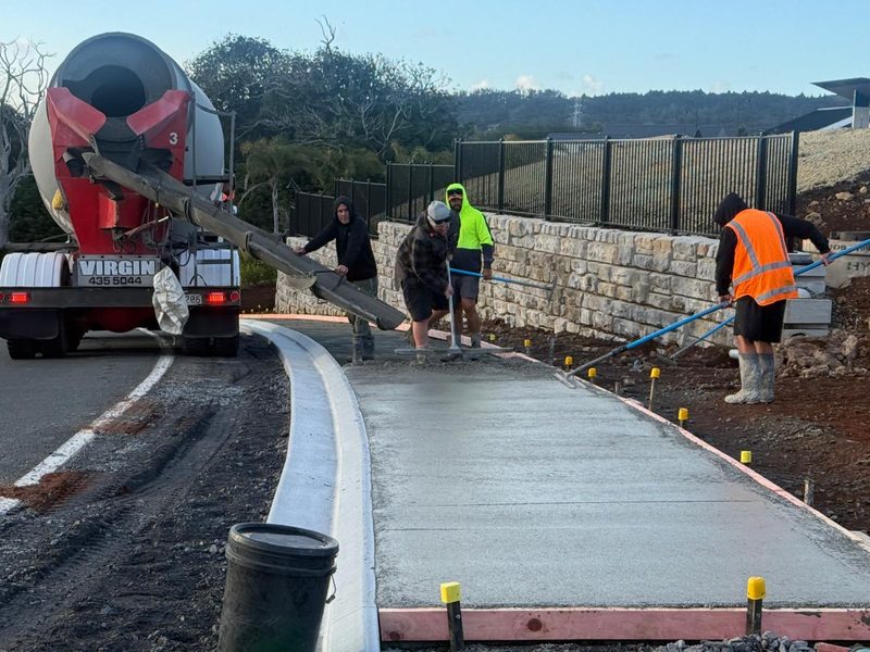 Crew pouring a concrete footpath beside new kerb and a stone block wall at Ashbury Heights, Maunu