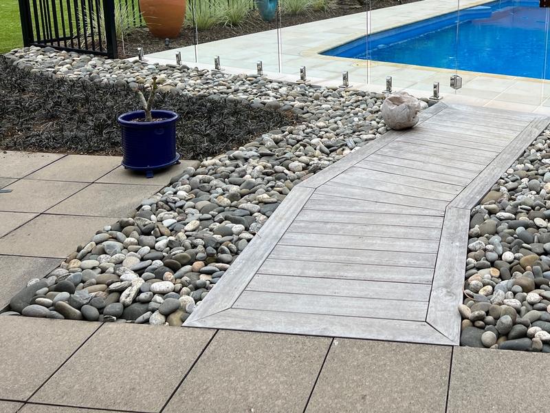 Timber walkway through river stones to paving and a glass-fenced pool